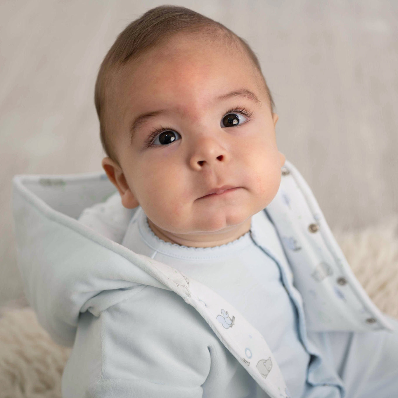 Handsome baby in a luxurious blue hooded sleepsuit, sitting on a fluffy blanket, expressing emotions